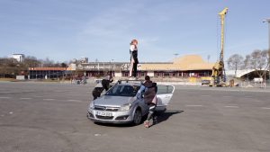 Eine Performerin steht auf dem Dach eines silbernen Autos auf einem großen leeren Platz und wird von zwei weiteren Performern geschoben.