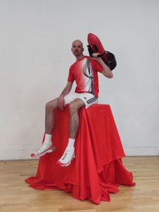 Künstler Niels Wehr sitzt in Sportkleidung auf einem hohen mit rotem Tuch bedeckten Podest.