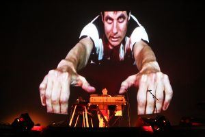 Felix Kubin auf einer Bühne mit seinem Musikpult, dahinter eine überdimensionierte Projektion von ihm, bei der er die Hände nach vorne ausstreckt.