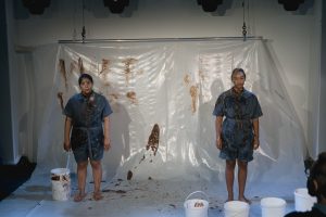 Zwei BPoC-Performerinnen stehen vor einer Plastikfolie, die mit dunklem Pudding beschmiert ist. Auch die Performerinnen sind mit diesem Pudding auf ihren Kleidern und in ihren Gesichtern beschmiert. Sie blicken zur Kamera.