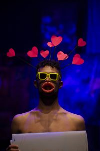 Ein junger Mann mit einer gelben Brille und pinken Herzen, die wie von seinen Haaren abstehen. Der Hintergrund ist blau.