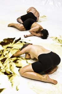 Drei Personen, oberkörperfrei, bewegen sich auf dem Boden. Der ist bedeckt von einer goldenen Flüssigkeit.