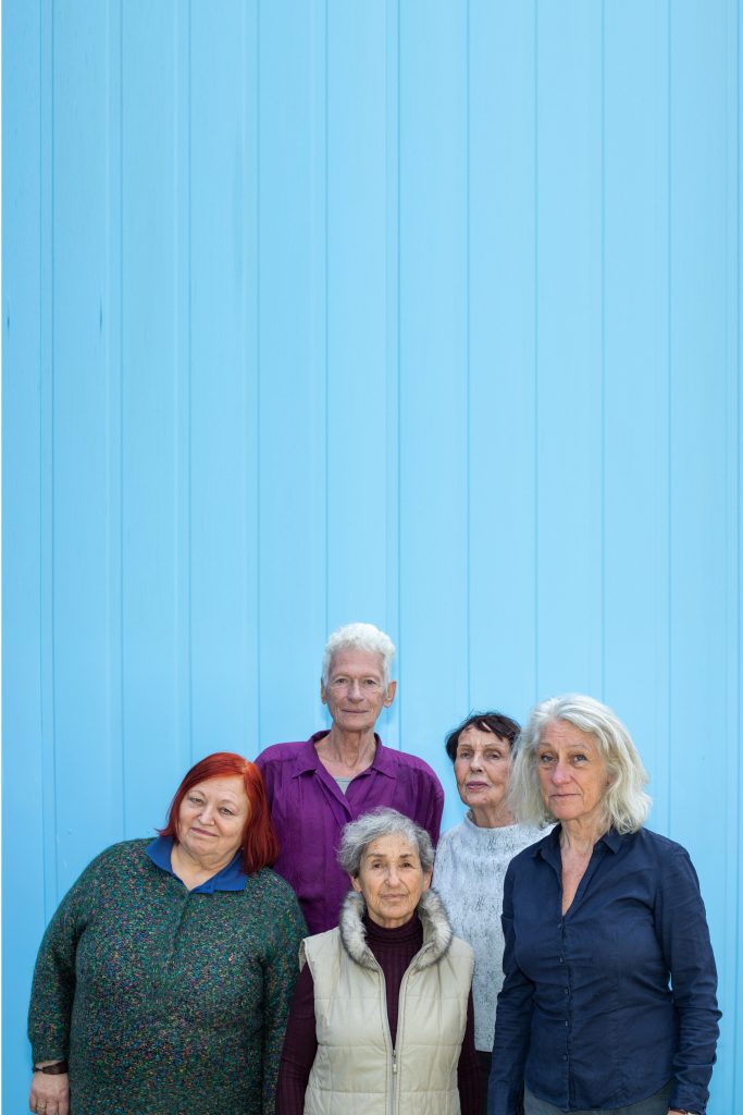 Personengruppe mit 5 Menschen über 70 Jahren in Frontalansicht bis zur Hälfte des Oberkörpers. Sie schauen in die Kamera. Sie stehen vor einer himmelblauen Wand, die die Hälfte des Bildes einnimmt.