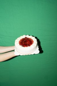 Auf dem Bild ist eine weiße Sahnetorte mit roter Erdberfüllung auf einem Papptablett zu sehen. Die Torte wird von zwei Händen gehalten, im Hintergrund sieht man ein grünes Stofftuch, welches das gesamte Bild ausfüllt.
