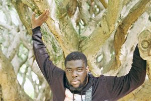 DJ Lycox steht vor einem Baum. Er trägt einen schwarzen Kapuzenpullover, grift mit erhobenen Händen an die Äste des Baumes und guckt in die Kamera.