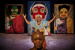 Eine Frau of Color in einem bunt gemusterten Gewand steht vor drei großen afrikanischen Gemälden und erhebt ihre Arme. Vor ihr thront eine afrikanische Büste.