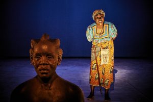 Eine Frau of Color in einem bunt gemusterten Gewand, die sich ans Publikum wendet. Vor ihr steht eine afrikanische Büste.