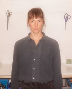 Porträt von DJ Carl. Sie steht, in dunklem Hemd gekleidet, vor einer Wand, an der zwei Scheren hängen.
