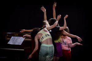Mehrere Performer*innen stehen hintereinander rechts neben einem Klavier und machen Moves mit ihren Armen.