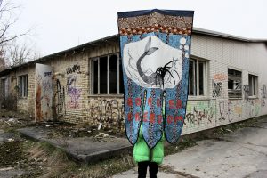 Das draußen aufgenommene Foto zeigt eine Person, die eine blau-gemusterte Fahne mit vor sich hochhält. Auf der Fahne steht: Border Freedom. Im Hintergrund steht ein verlassenes Gebäude mit viel Graffiti an den Wänden.
