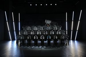 In einem Bühnenraum mit einer Art kleinen Tribüne aus Plastikstühlen steht eine Performerin in der hintersten den Stuhlreihe und hebt einen der Stühle hoch.