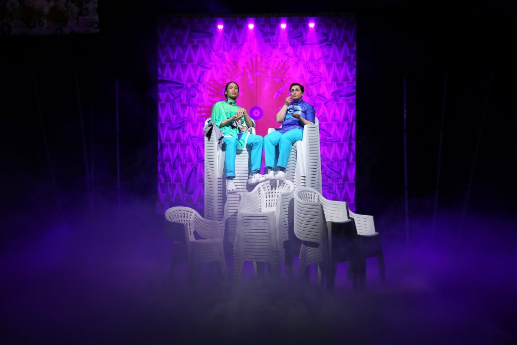 Zwei Performer*innen in bunter Kleidung sitzen auf einer Vielzahl von übereinander gestapelten weißen Plastikstühlen und sind dabei etwas zu essen. Im Hintergrund ist eine violett illuminierter Vorhang, in dessen Mitte ein Kreis von Handschatten abgebildet ist.