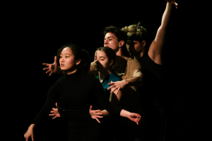 Vier Performer*innen stehen hintereinander vor einem schwarzen Hintergrund und führen unterschiedliche Handbewegungen durch.
