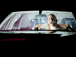 Eine Performerin sitzt in einem beleuchteten Auto und schreit, ihre Hände sind erhoben. Außerhalb des Autos ist es dunkel.