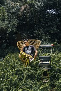 Ein vietnamesischer Künstler sitzt in einem Korbstuhl inmitten von Pflanzen. Neben ihm steht ein Radio, dahinter ein Mischpult.