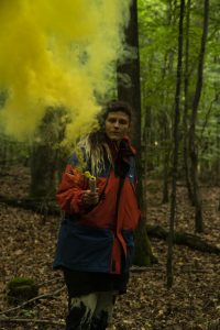 Eine Frau mit schwarz-roter Jacke steht im Wald und zündet eine Rauchbombe.
