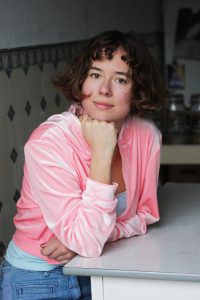 Porträt der Comiczeichnerin und Autorin Helena Baumeister. Sie sitzt an einem Küchentisch, hat den Kopf auf der Hand abgestützt und blickt direkt in die Kamera.