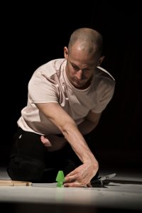 Der Performer Michael Turinsky kniet auf dem Boden und verschränkt seinen linken Arm. Er blickt auf einen Miniaturbaum aus Holz.