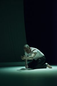 Turinsky kniet in einem dunkelgrün beleuchteten Raum und streckt seinen rechten Arm nach vorne aus.
