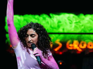 Die Performerin Nastaran Razawi Khorasani steht im Vordergrund des Bildes. Sie hat lange dunkle Locken, ihre Arme sind mit pinker Farbe bemalt. Sie hält ein Mikrofon und streckt den anderen Arm hoch in die Luft. Im Hintergrund orangefarbene Schrift unter einer grellen grünen Farbfläche. Die Schrift ist verschwommen und nicht lesbar.