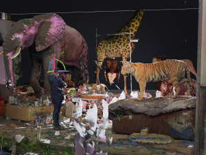 Ein computergeneriertes Bild von ausgestopften Tieren: Ein Elefant, eine Giraffe, ein Tiger und im Hintergrund ein Löwe. Auf dem Boden sind eine Menge unerkennbarer Gegenstände ins Bild eingefügt, wobei teilweise unterschiedlich farbige grobe Bildpixel erkennbar sind. Inmitten der Tiere ist das verpixelte Bild eines Menschen eingefügt.