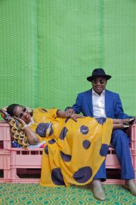 Die Musiker*innen Amadou und Mariam sitzen und liegen auf einer Couch aus rosafarbenen Plastikkisten. Der Hintergrund besteht aus grüner Plane. Mariam trägt ein gelbes Kleid mit großen blauen Punkten. Amadou trägt einen blauen Anzug, ein weißes Hemd und einen schwarzen Hut.