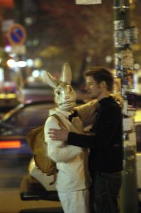 Eine Foto von einer Straßenszene, verwackelt aufgenommen. Im Vordergrund umarmen sich ein Mann und eine als Hase verkleidete Person und blicken in Richtung Kamera.