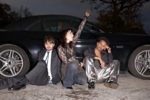 Auf dem Bild sitzen die drei Künstler*innen der Gruppe Sharktank vor einem Auto. Zwei von ihnen gucken in die Kamera und eine macht ein Selfie mit einer Digitalkamera. Hinter dem Auto stehen Bäume vor einem grauen Abendhimmel.