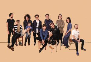 Auf dem Bild befinden sich die 11 Mitglieder von der Band Gorilla Club vor einem orangenen Hintergrund. Manche von ihnen sitzen auf Holzstühlen, während andere stehen.
