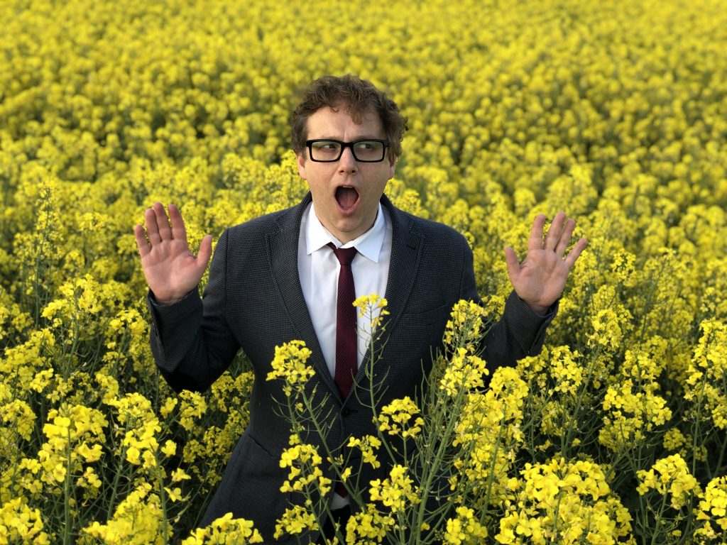 Auf dem Foto steht der Sänger Sven van Thom in einem gelben Rapsfeld. Er trägt einen Anzug und eine Brille. Sein Mund ist geöffnet und er hält die Hände hoch.