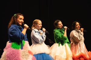 Eine Foto von den vier Performerinnen auf der Bühne in Großaufnahme. Sie tragen bunte Kleider mit wallenden Röcken und halten jeweils ein Mikrofon vor ihren Mund.