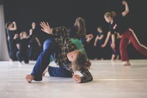 Das Foto zeigt mehrere tanzende Personen in einem Proberaum. Im Vordergrund ist die Tänzerin Jerneja Fekonja in einer Bewegung am Boden, bei der sie das rechte Bein nach vorne und den rechten Arm nach oben streckt. Sie trägt ein gelb-schwarz kariertes Oberteil und eine Blaue Hose.