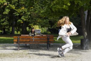 Das Foto ist in einem Park mit grünen Bäumen aufgenommen und zeigt die Tänzerin Jerneja Fekonja mitten in einer dynamischen Bewegung. Ihre langen hellen Haare wehen zur Seite. Sie trägt einen weißen Blazer.