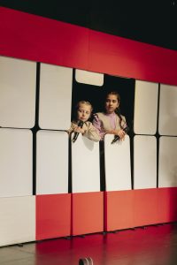 Auf dem Foto stehen zwei Mädchen hinter einer Wand aus roten und weißen Platten, die auf einer Bühne aufgebaut ist. Sie gucken durch eine Öffnung in der Wand hindurch schräg Richtung Publikum und halten Mikrofone in der Hand.