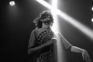 Auf dem schwarz-weiß-Foto steht der Künstler Ryskinder auf einer Bühne und hält ein Mikrofon in der Hand. Das Licht einer der Scheinwerfer streift ihn und er hat seine Augen geschlossen.