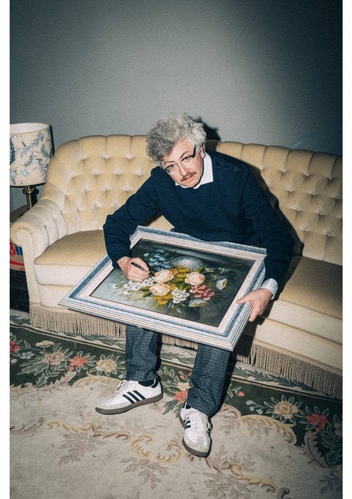Auf dem Foto in Retrooptik sitzt der Autor Gereon Klug auf einem altmodischen Sofa in einem altmodisch eingerichteten Raum. Er hält ein Stillleben mit Blumen in den Händen und schreibt etwas darauf.