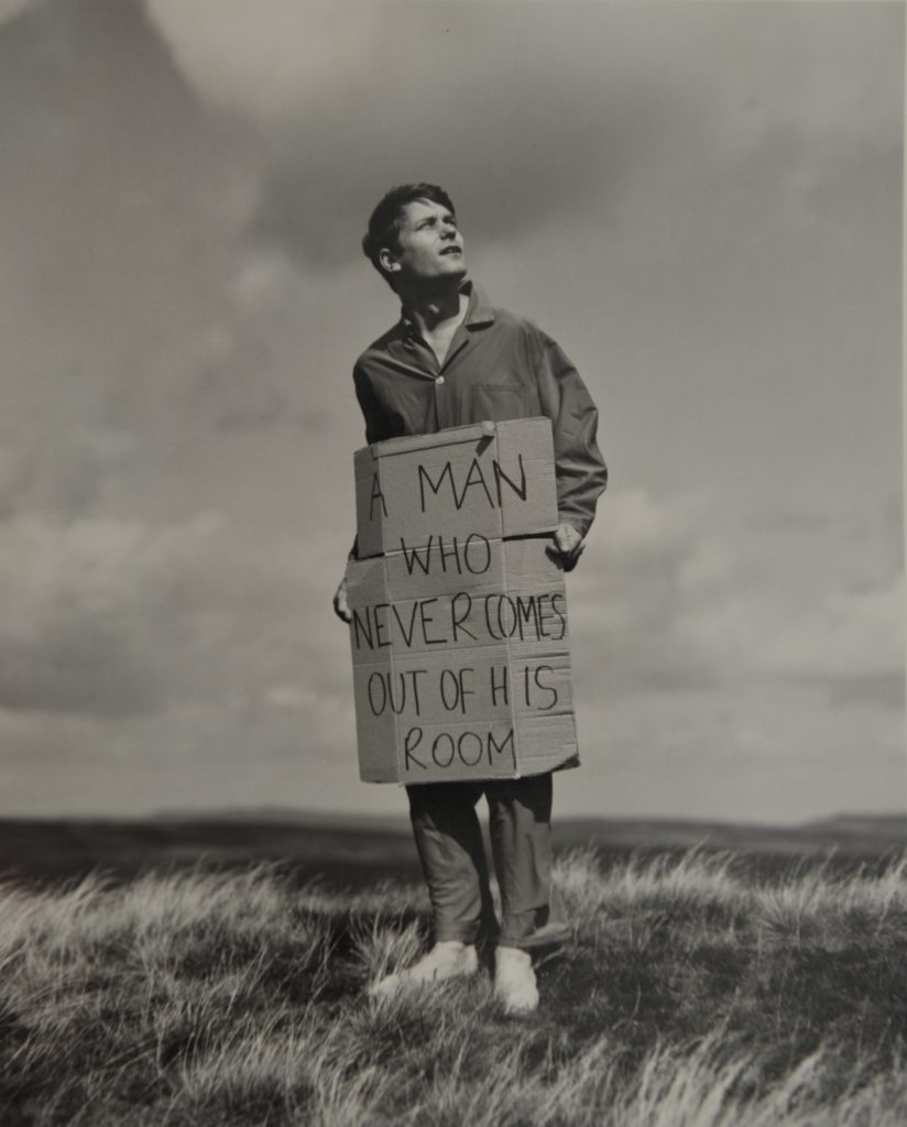 Ein Schwarz-weiß-Foto von einem Mann, der in einer hügeligen Landschaft steht. Er hält ein Schild mit der Aufschrift „A man who never comes out of his room“ in den Händen.