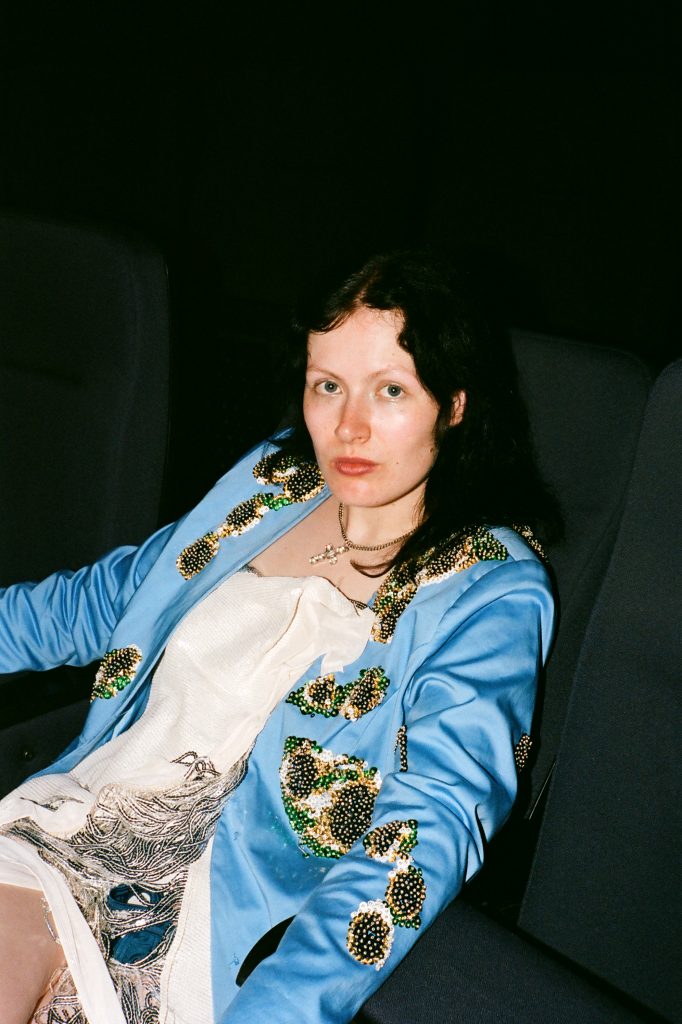 Auf dem Bild sitzt die Musikerin Rosa Anschütz auf einem grauen Sessel und guckt in Richtung Kamera. Sie trägt eine blaue Jacke. Der Hintergrund ist dunkel.