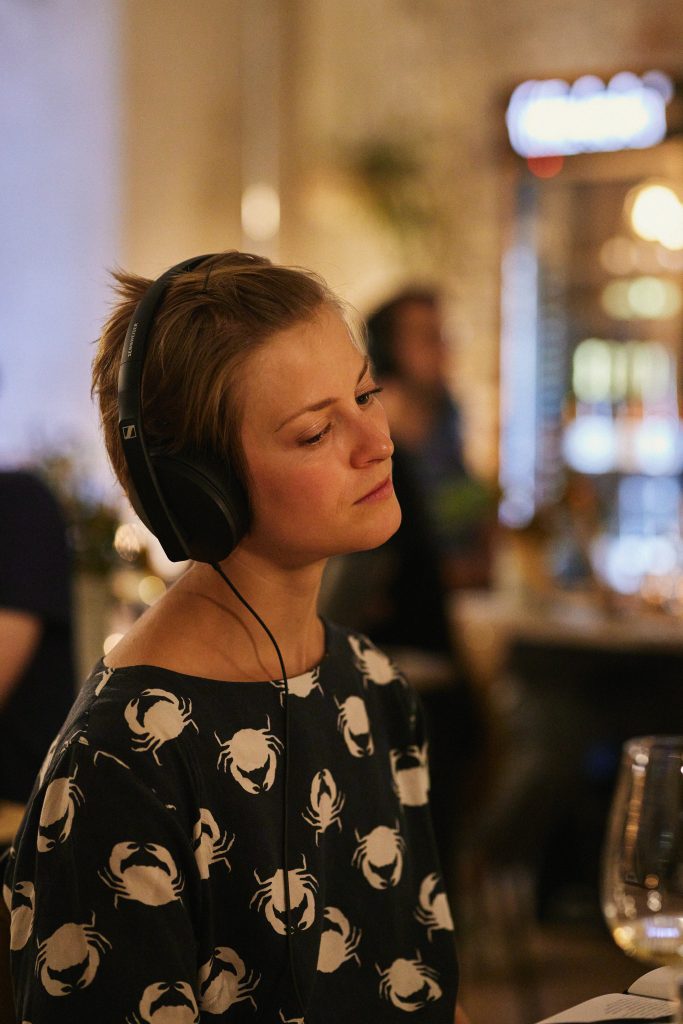 Auf dem Foto sitzt eine junge Frau mit kurzen Haaren in einem Café. Sie hat große Kopfhörer auf.